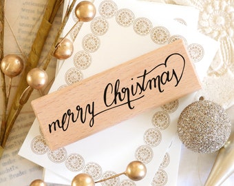 May include: A wooden stamp with the words "merry Christmas" in elegant black script. The stamp is set against a backdrop of decorative cards, gold ornaments, and floral accents, creating a festive holiday theme.