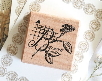 May include: A wooden stamp with the word "BELIEVE" in a decorative font, accompanied by floral and grid designs. The stamp is on a white surface with a pattern of gold circular designs.