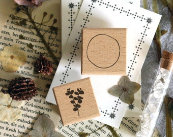 May include: Two wooden stamps with botanical designs and a circle, alongside dried flowers and a glass vial filled with crystals. The stamps are on a vintage paper background, creating a rustic, artistic aesthetic.