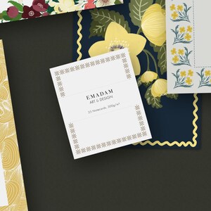 May include: Stationery set featuring notecards and floral designs. The main notecard displays "EMADAM ART & DESIGN" and "25 Notecards, 300g/m²". The background includes floral patterns in yellow, white, and dark blue, with a gold border.