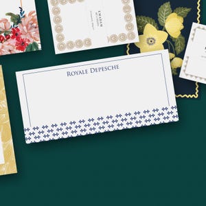 Può includere: Carta bianca con la scritta "ROYA LE DEPESCHE" in blu navy. Il bordo inferiore presenta un motivo floreale blu navy. Sono visibili altri articoli di cancelleria con disegni floreali e dorati.