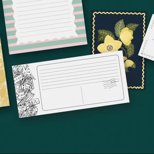 Può includere: Una collezione di articoli di cancelleria decorativi, tra cui cartoline e carta da lettere, disposti su una superficie verde scuro. Le cartoline presentano disegni floreali e spazi a righe per la scrittura. Una cartolina ha un francobollo aereo vintage.