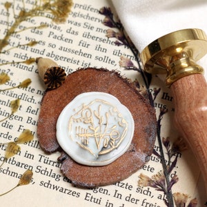 May include: A vintage-style wax seal stamp with a wooden handle and a brass stamp head. The seal features a white wax impression with gold-colored botanical design. The stamp rests on a piece of wood, with dried flowers and a book in the background.