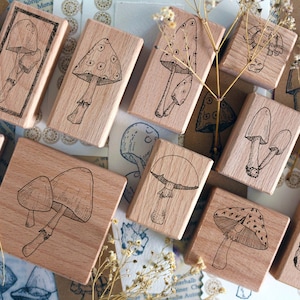 Op de afbeelding: Een verzameling houten stempels met paddenstoelontwerpen. Elke stempel heeft een illustratie van een paddenstoel in zwarte inkt. De stempels zijn gerangschikt op een oppervlak met gedroogde bloemen en papier.