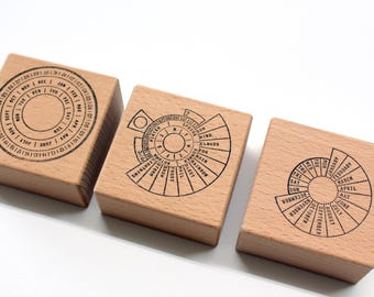 May include: Three wooden stamps with black ink designs. The stamps feature circular and semi-circular calendar layouts with months, days, and weather conditions. The stamps are made of light-colored wood.