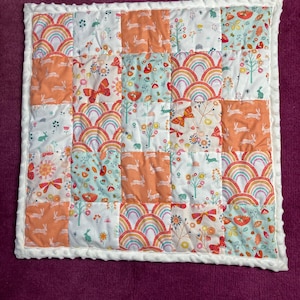 May include: A patchwork quilt with squares displaying rainbows, butterflies, and bunnies in orange, teal, and white. The quilt has a white, textured border, suitable for a baby or child.