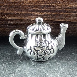 May include: A silver teapot charm featuring a floral design. The teapot has a lid and a spout. The charm is small and intricate.