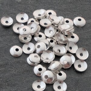 Può includere: Una collezione di piccole perline metalliche circolari color argento con una forma concava a scodella e un singolo foro centrale. Questi componenti metallici presentano una superficie liscia e riflettente e sono sparsi in un mucchio.
