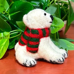May include: A white felt polar bear wearing a red and green striped scarf. The bear is sitting on a red surface.