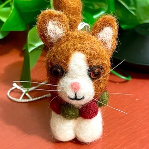 May include: A small, felted cat ornament with a white face, brown fur, and a green and red pom-pom scarf.
