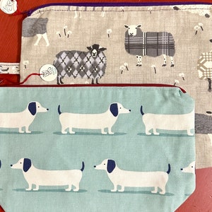 May include: Two fabric zippered pouches. The top pouch is beige with a pattern of black and white sheep. The bottom pouch is teal with a pattern of white dachshunds.