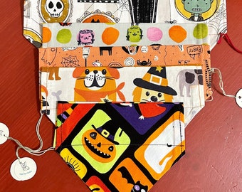 May include: A stack of five colorful Halloween-themed dog bandanas. The bandanas feature various designs, including pumpkins, ghosts, bats, and witches. The bandanas are made of fabric and have a sewn edge.