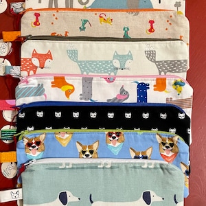May include: A stack of nine colorful zippered pouches with various animal prints. The pouches are made of fabric and have a zipper closure. The prints include elephants, foxes, dogs, cats, and corgis.