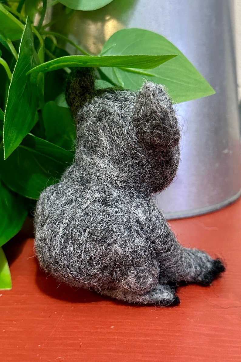May include: A grey felt koala sitting on a red surface. The koala is looking to the right and has its arms raised.