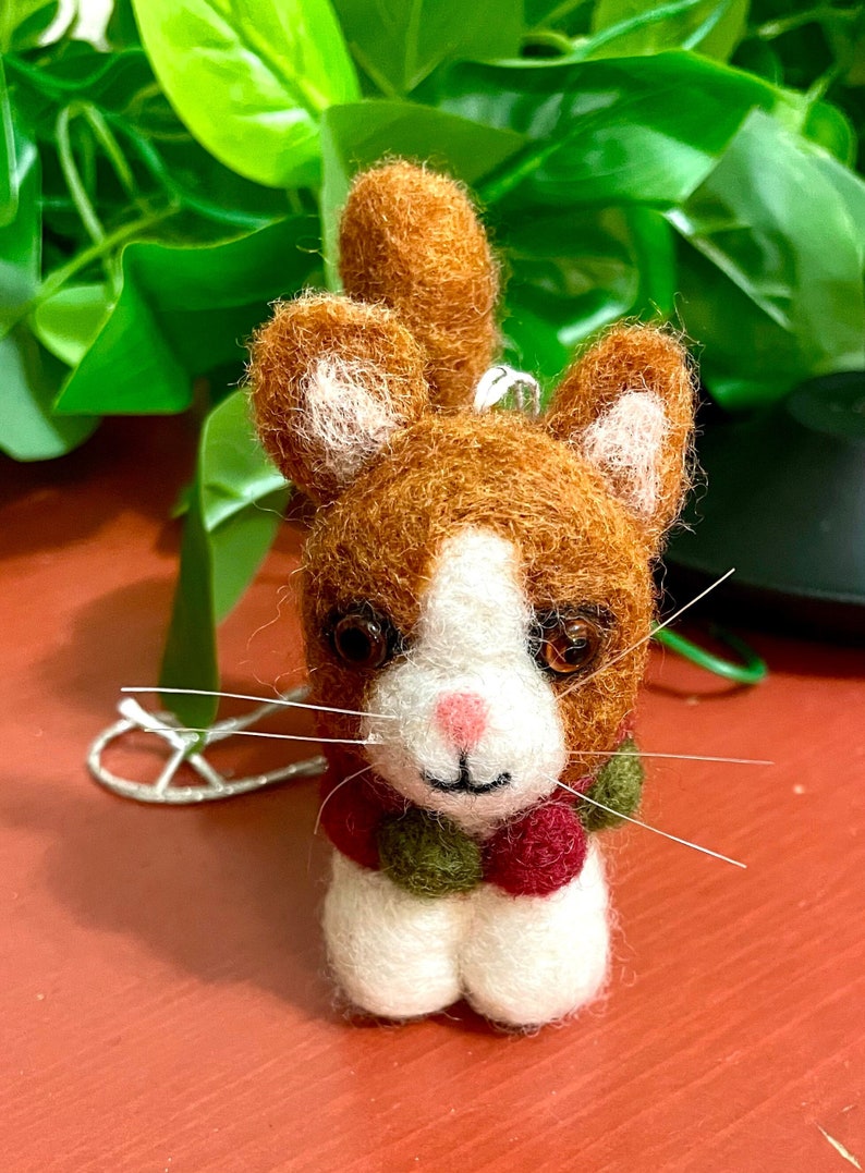May include: A small, felted cat ornament with a white face, brown fur, and a green and red pom-pom scarf.