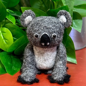 May include: A grey and white felt koala toy with black eyes and a black nose. The koala is sitting on a red surface.