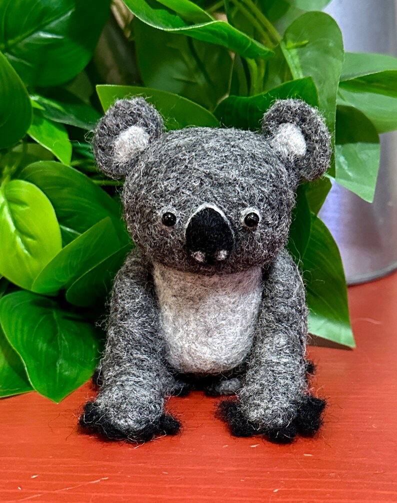 May include: A grey and white felt koala toy with black eyes and a black nose. The koala is sitting on a red surface.