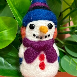 May include: A small, white felt snowman wearing a blue and purple hat and a purple scarf. The snowman has black eyes, a small orange nose, and a red button.