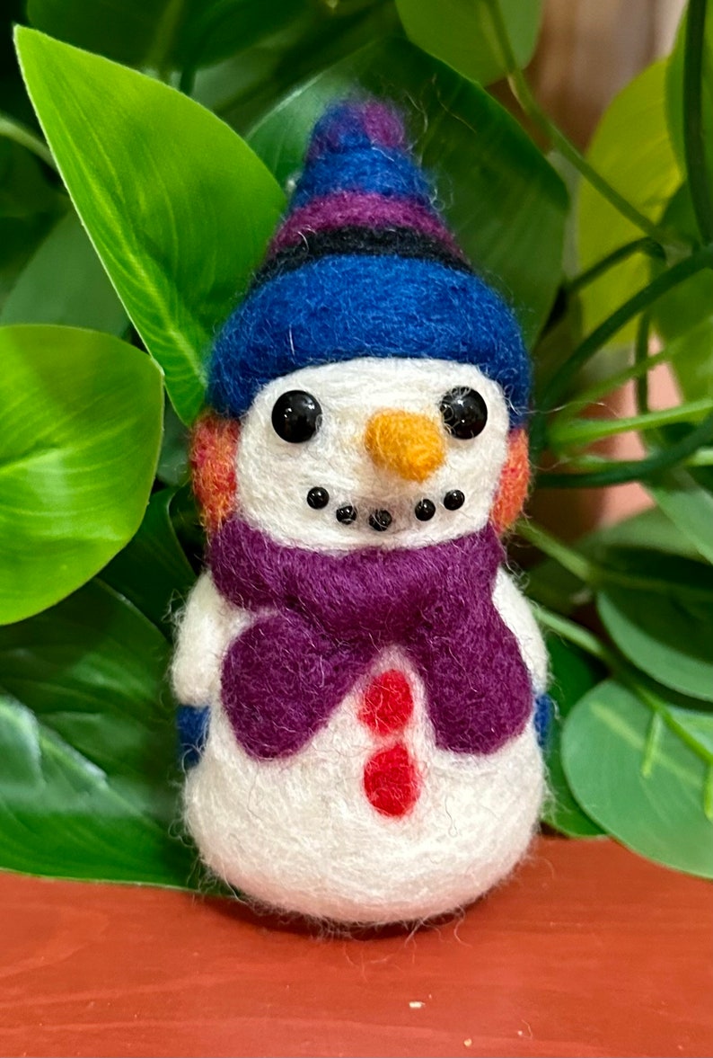 May include: A small, white felt snowman wearing a blue and purple hat and a purple scarf. The snowman has black eyes, a small orange nose, and a red button.