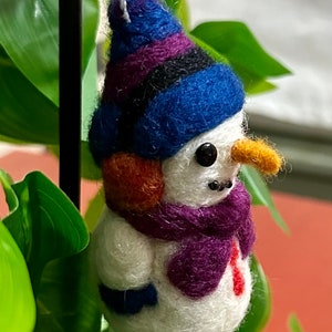 May include: A small felt snowman ornament with a blue, purple and black striped hat, an orange ear, a purple scarf and a white body.