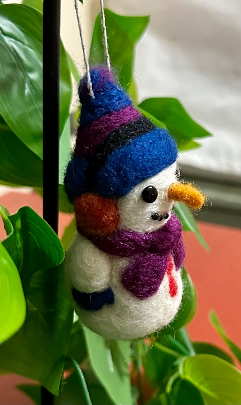 May include: A small felt snowman ornament with a blue, purple and black striped hat, an orange ear, a purple scarf and a white body.