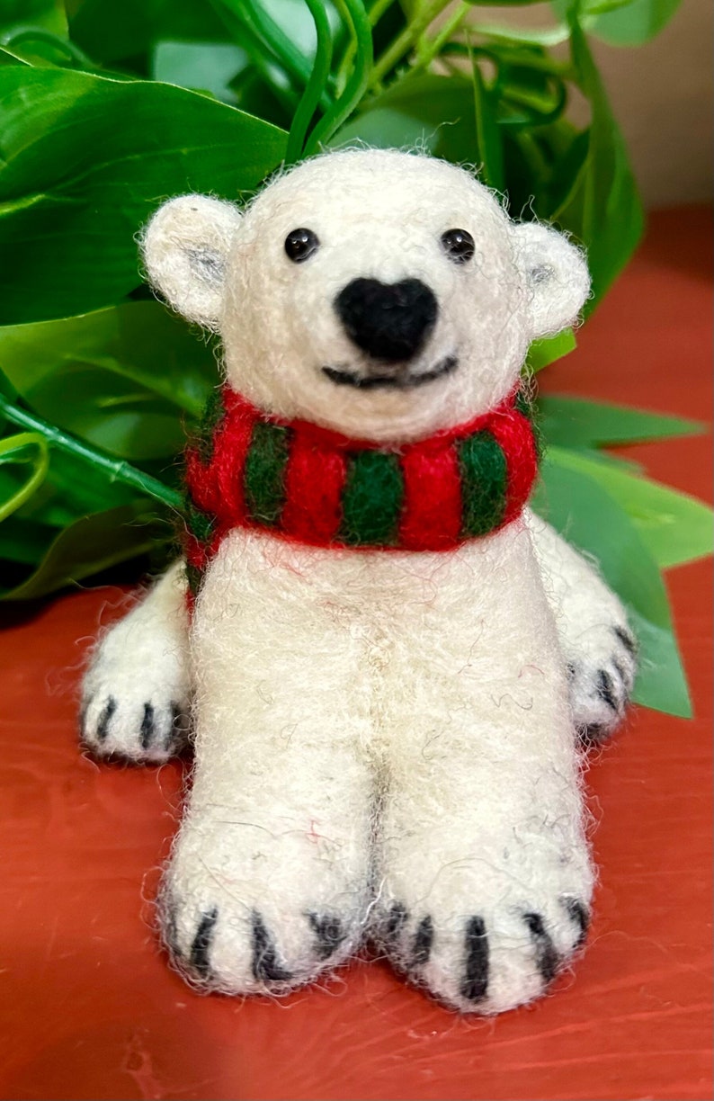 May include: A white felt polar bear wearing a red and green striped scarf. The bear is sitting with its paws in front of it.