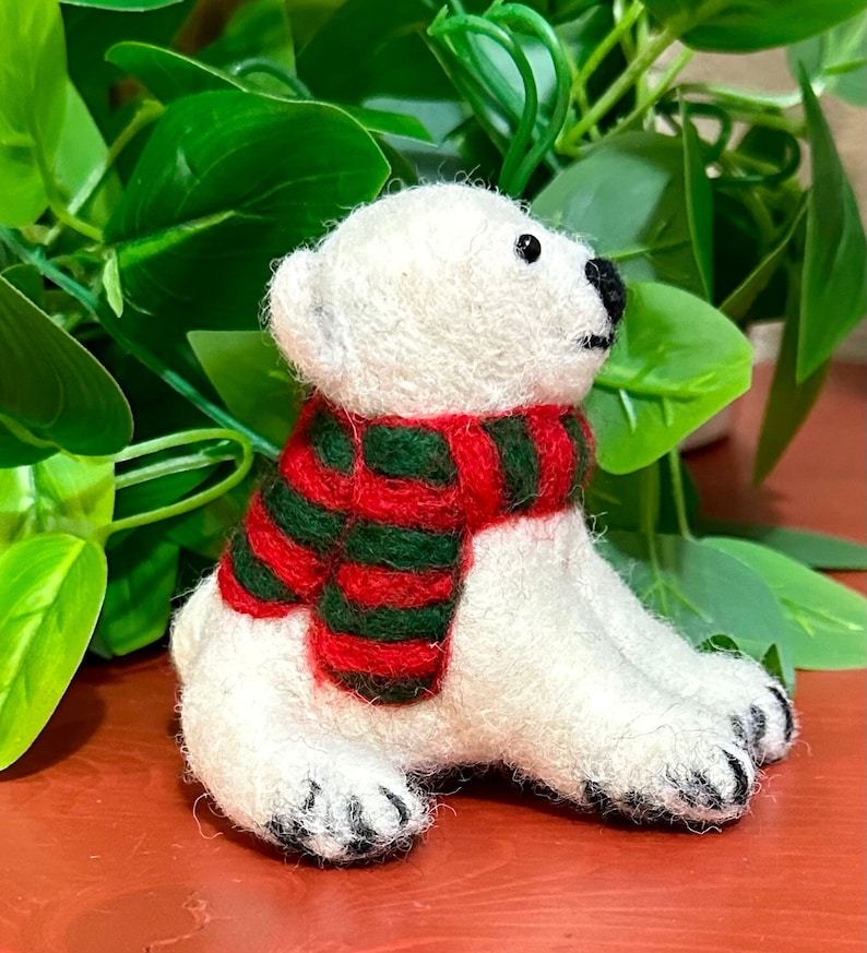 May include: A white felt polar bear wearing a red and green striped scarf. The bear is sitting on a red surface.