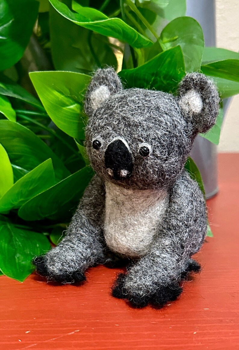 May include: A grey and white felt koala sitting on a red surface. The koala has black eyes and a black nose.