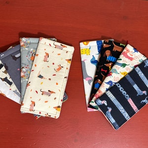 May include: A set of eight fabric eyeglass cases featuring different dachshund designs. The cases are made of a soft, quilted fabric and have a zipper closure. The designs include dachshunds in various colours and patterns, including black, white, brown, and grey.