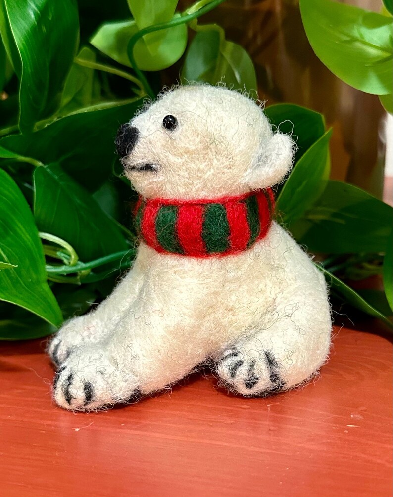 May include: A white felt polar bear ornament wearing a red and green striped scarf. The bear is sitting with its paws tucked under its body.