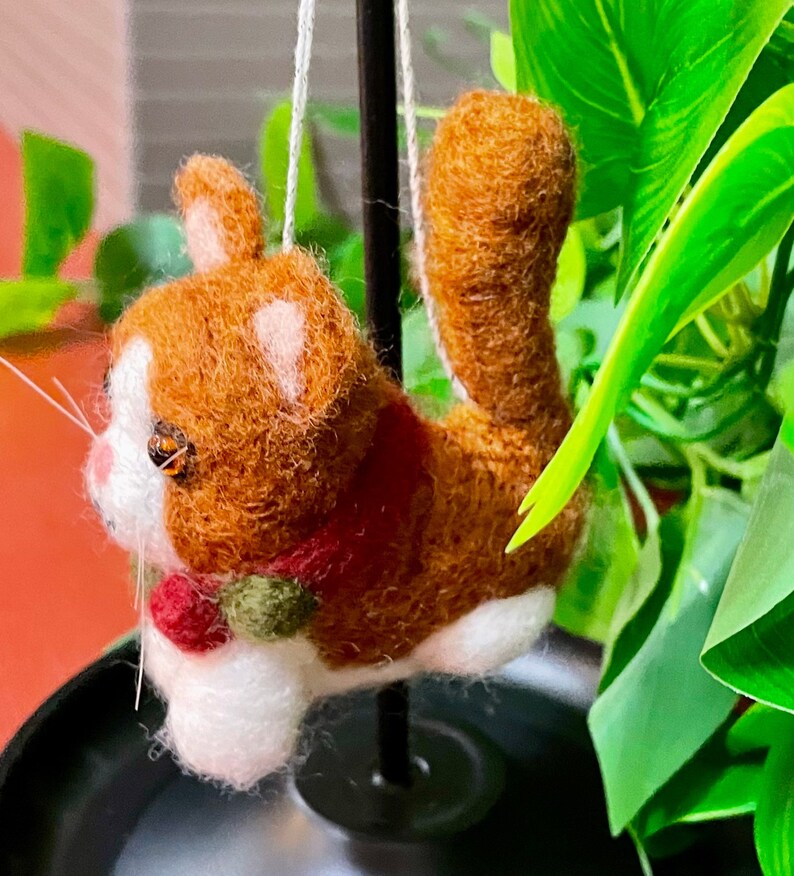 May include: A brown and white felted cat ornament with a red and green scarf. The cat is hanging from a black stand.