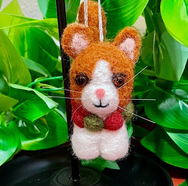 May include: A felt cat ornament with a white face, orange and white fur, and a red and green scarf. The cat has a pink nose and brown eyes.
