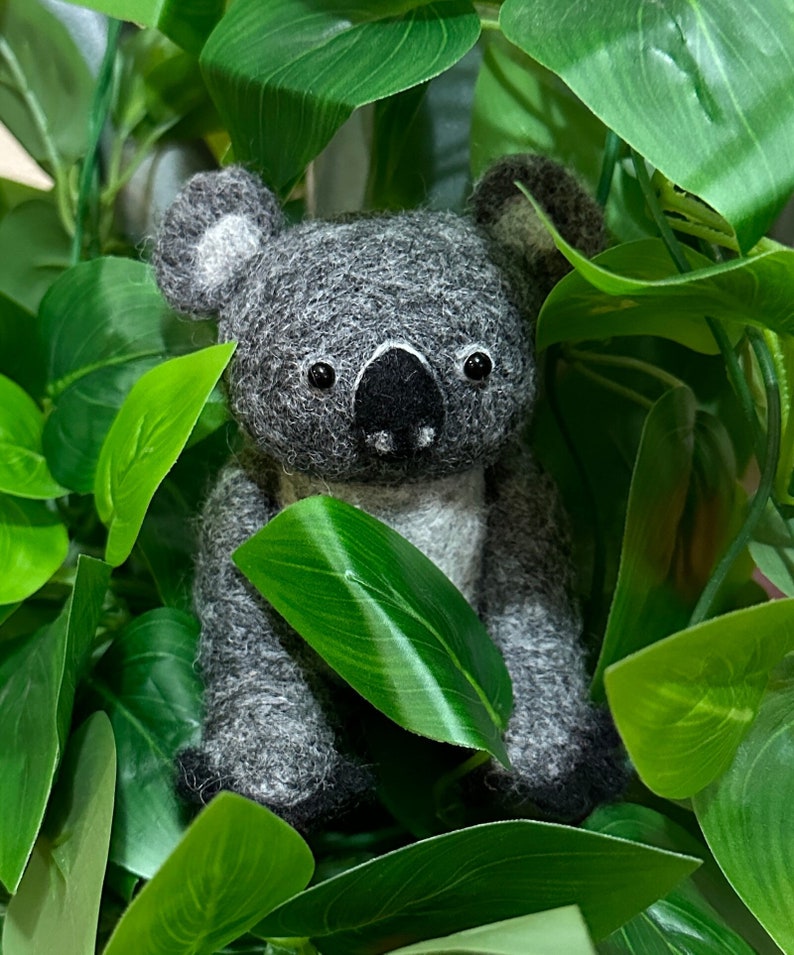 May include: A grey and white koala plush toy is nestled amongst green leaves. The koala has a black nose and small black eyes.