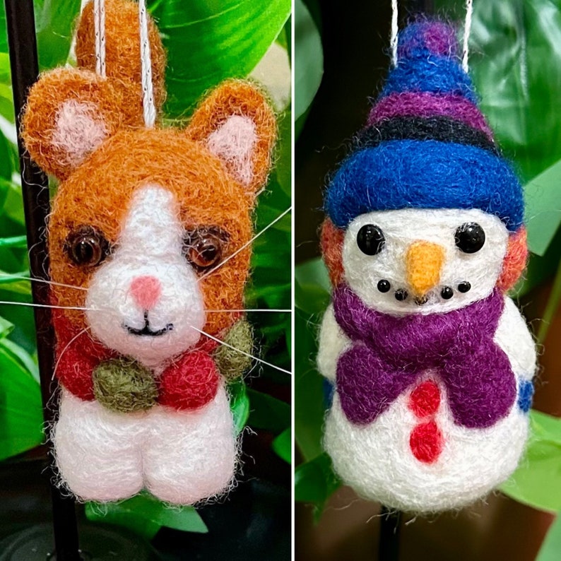 May include: Two felt ornaments, a brown and white cat and a white snowman with a purple hat and scarf. The cat has a red and green scarf around its neck.