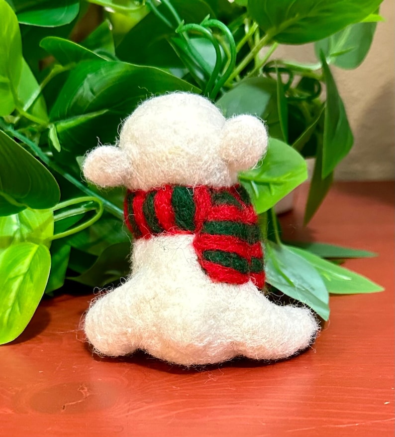 May include: A white felt animal wearing a red and green striped scarf. The animal is sitting on a red surface.