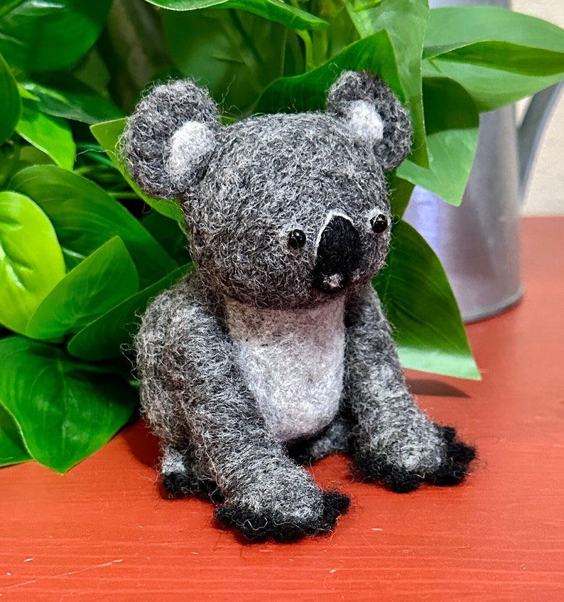 May include: A grey and white felt koala plush toy with black eyes and a black nose. The koala is sitting on a red surface.