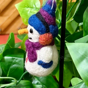 May include: A felt snowman ornament with a purple and blue hat, orange nose, and purple scarf. The ornament is hanging from a black string.