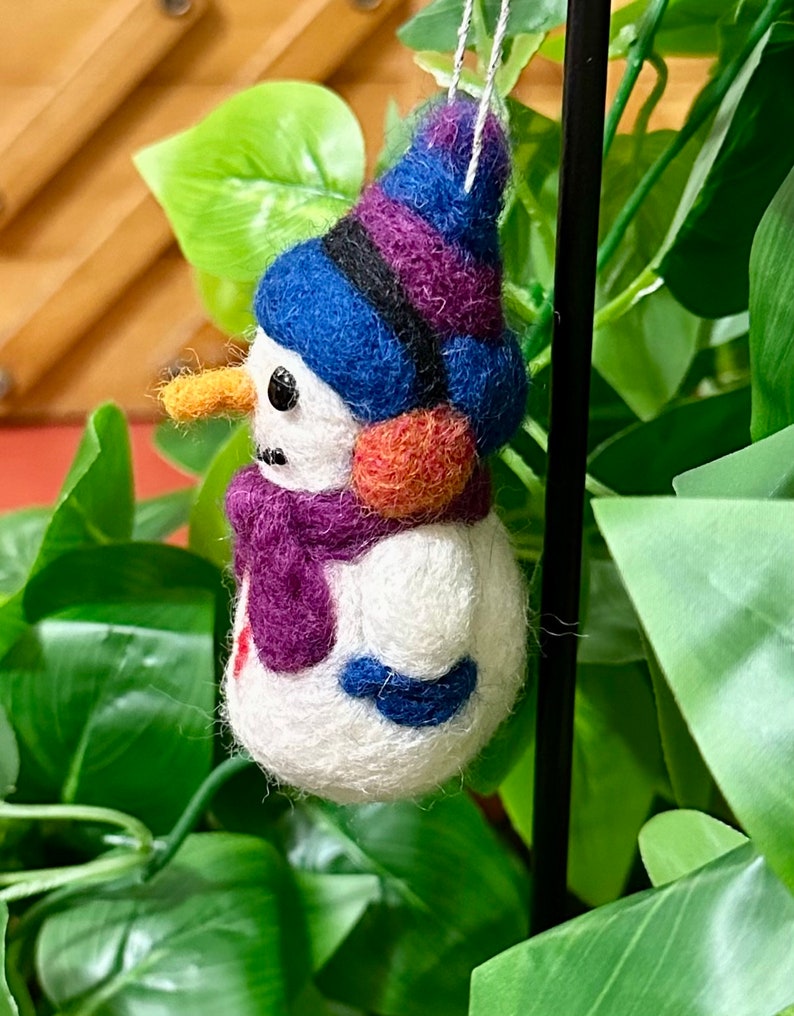 May include: A felt snowman ornament with a purple and blue hat, orange nose, and purple scarf. The ornament is hanging from a black string.