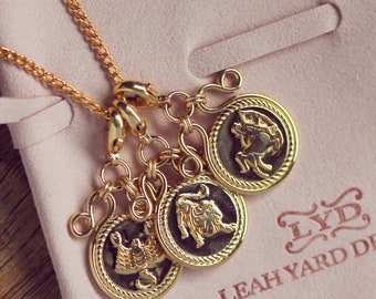 May include: Gold necklace with four charms featuring zodiac signs. The charms are attached to the chain with small gold rings. The charms are round and have a raised edge. The zodiac signs are Sagittarius, Leo, Scorpio, and Capricorn. The necklace is on a pink background with the text "LYD, LEAH YARD DE" in gold.