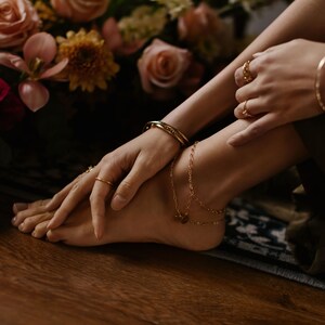 May include: A woman's ankle with a gold chain anklet with a small charm. The anklet is layered with a second, thinner chain. The woman is wearing a gold bracelet on her wrist and a gold ring on her finger.