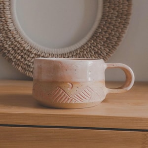 Op de afbeelding: Handgemaakte keramische mok met een lichtroze en beige kleurenschema. Het bovenste gedeelte is roze, terwijl het onderste gedeelte een beige, getextureerd ontwerp heeft. De mok heeft een afgerond handvat en wordt waarschijnlijk gebruikt voor dranken.