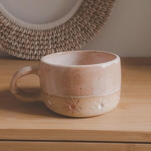 Puede incluir: Taza de cerámica hecha a mano con una parte superior rosa claro y una parte inferior beige. La parte inferior presenta un diseño de sol repetido. La taza tiene forma redondeada y un asa curva. La taza está sobre una superficie de madera.