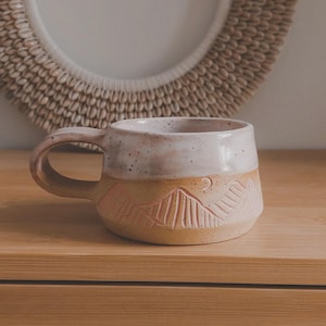 Op de afbeelding: Een handgemaakte keramische mok met een bergontwerp. De mok heeft een beige basis met een uitgesneden bergketen en een crèmekleurige bovenkant. Het handvat is gebogen en de mok is ongeveer 10 cm hoog.