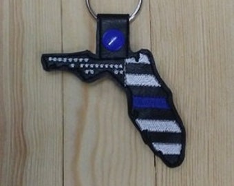 Puede incluir: Un llavero de cuero negro con forma del estado de Florida con un diseño de línea azul fina azul y blanco.
