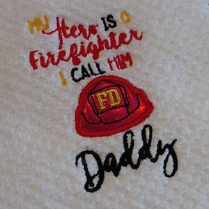 Puede incluir: Toalla de cocina blanca bordada con el texto "Mi héroe es un bombero Lo llamo Papá" y un gráfico de casco de bombero rojo y amarillo.