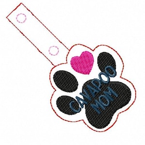 May include: A black and white paw print with a pink heart and the text "Cavapoo Mom" in blue thread. The paw print is attached to a white strap with two red circles.