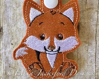 Könnte beinhalten: Ein brauner und weißer Fuchs-Schlüsselanhänger mit glitzernder Oberfläche. Der Fuchs sitzt mit seinen Pfoten zusammen und hat eine weiße Schnauze und Ohren.