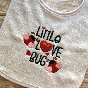 Puede incluir: Babero blanco para bebé con mariquitas bordadas en rojo y negro y el texto "Little Love Bug" en negro.