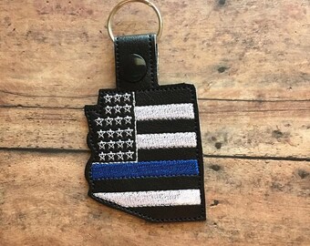 Puede incluir: Llavero de cuero negro con un diseño de bandera estadounidense de línea azul delgada. El llavero tiene un anillo de metal plateado.