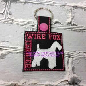 Könnte beinhalten: Schwarzer Leder-Schlüsselanhänger mit rosa Nähten und einem rosa Knopf. Der Schlüsselanhänger zeigt eine weiße Silhouette eines Wire Fox Terriers mit dem Text "WIRE FOX TERRIER" in rosa Nähten.